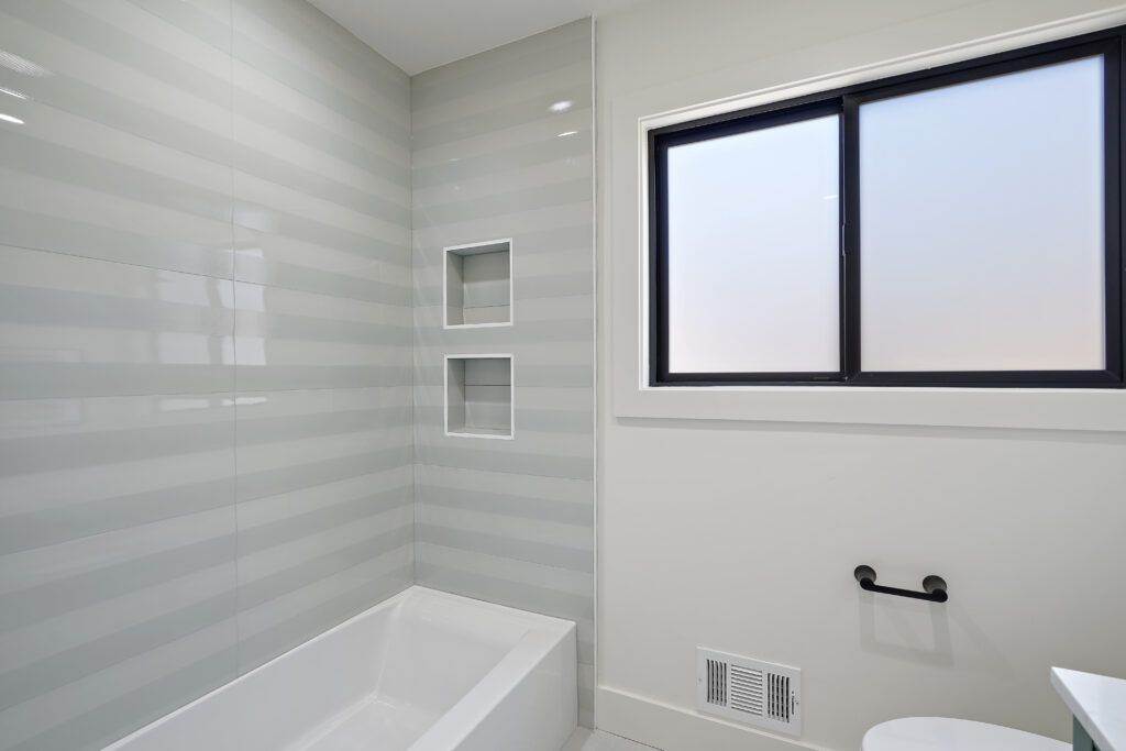 Modern bathroom interior featuring a built-in bathtub, gray tiled walls with shelving, and a large window with a black frame, completed in the post-construction phase.