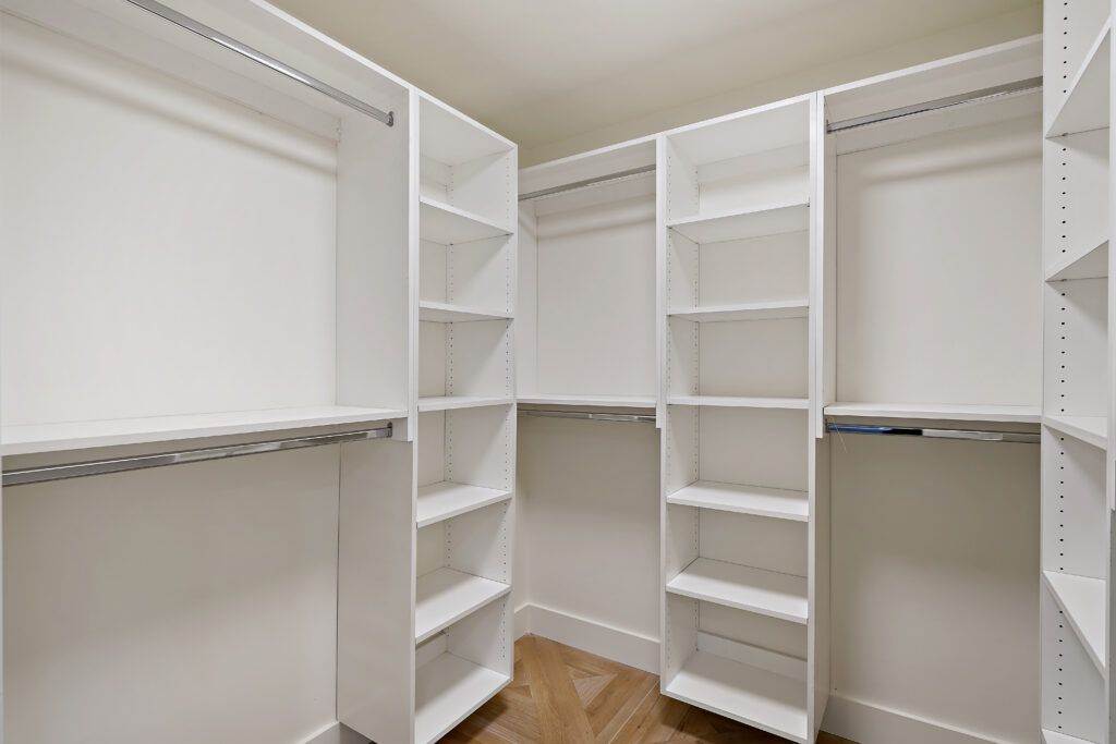 An empty post-construction walk-in closet with white shelves and hanging rods on three walls, featuring a light wooden floor.