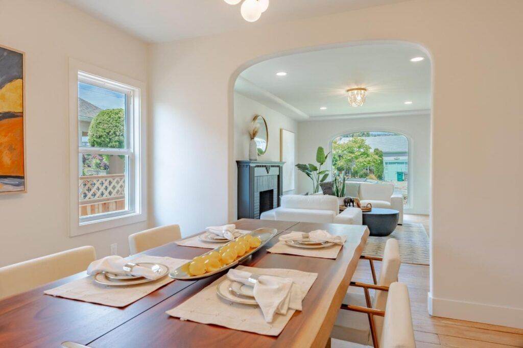 Modern dining room with a wooden table set for six, leading to an archway with a view into a living area. Bright, natural light fills the space, highlighting the post-construction finish.