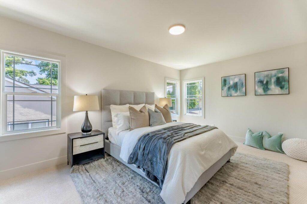 Bright, modern bedroom with a large bed, white and gray bedding, a bedside table with a lamp, and abstract wall art above the bed. This room is ideal for showcasing in a construction portfolio.