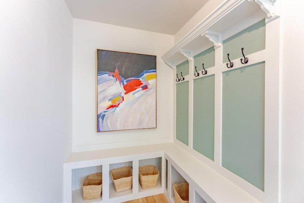 A tidy post-construction entryway with built-in seating and storage, light green paneling, white hooks for hanging items, and a colorful abstract painting on the wall.