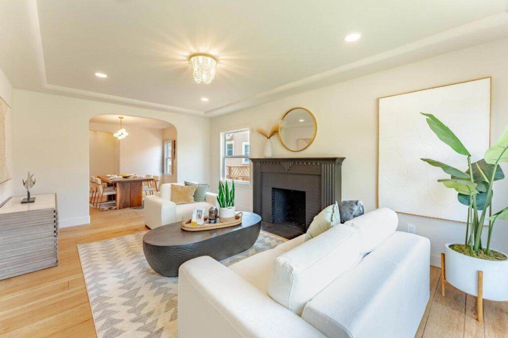 Bright, spacious living room with white walls, a dark fireplace, modern furniture, and large plants by a sunny window, leading through an archway to a dining area in a post-construction portfolio.