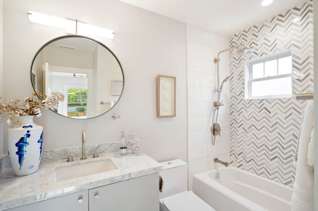 Modern bathroom with a circular mirror, white marble countertop, patterned wall, and a bathtub, featured in our post-construction portfolio.