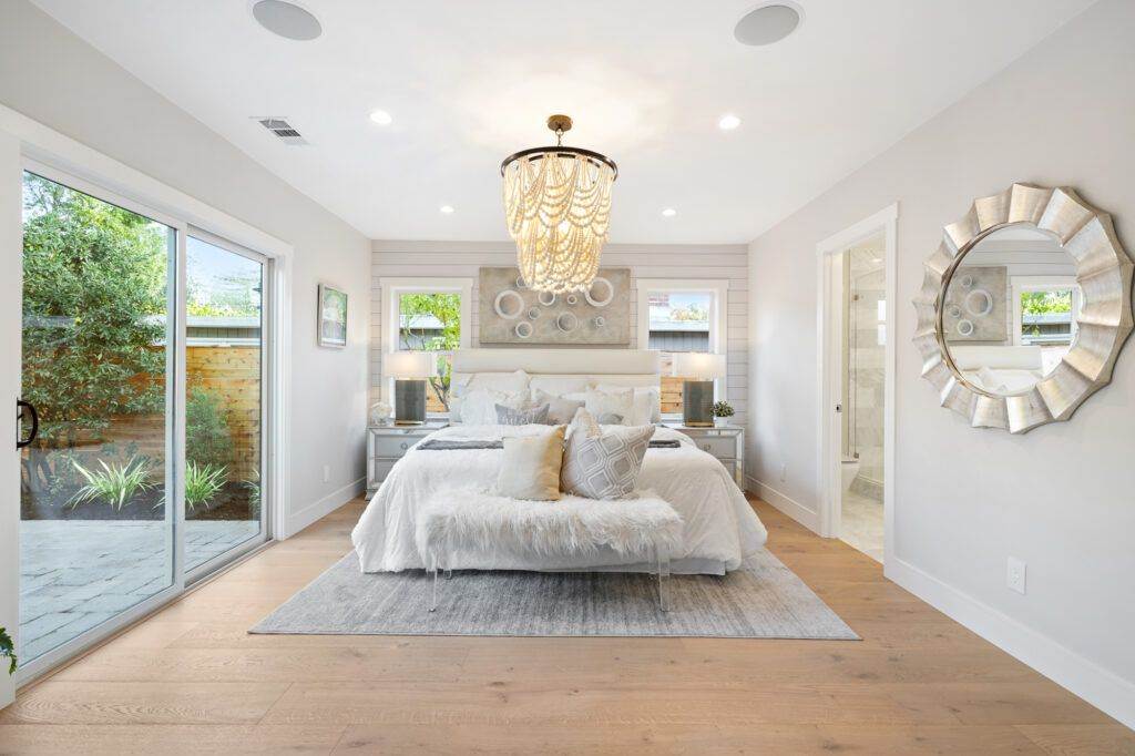 Bright, modern bedroom with a large bed, chandelier, glass doors leading to a garden, and decorative mirrors on the walls newly renovated in our post-construction portfolio.