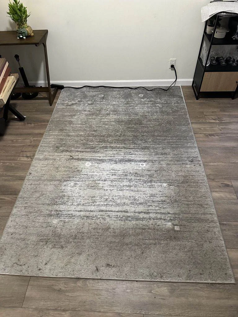A worn gray area rug on a wooden floor in a room with an upholstered desk and a shelving unit in the background.