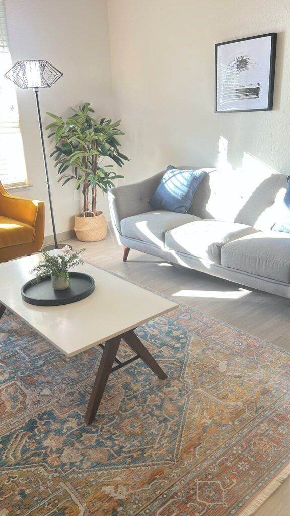 A bright living room with a gray sofa, yellow armchair, a white coffee table on an ornate rug, a potted plant, and wall art, bathed in natural sunlight after a Master