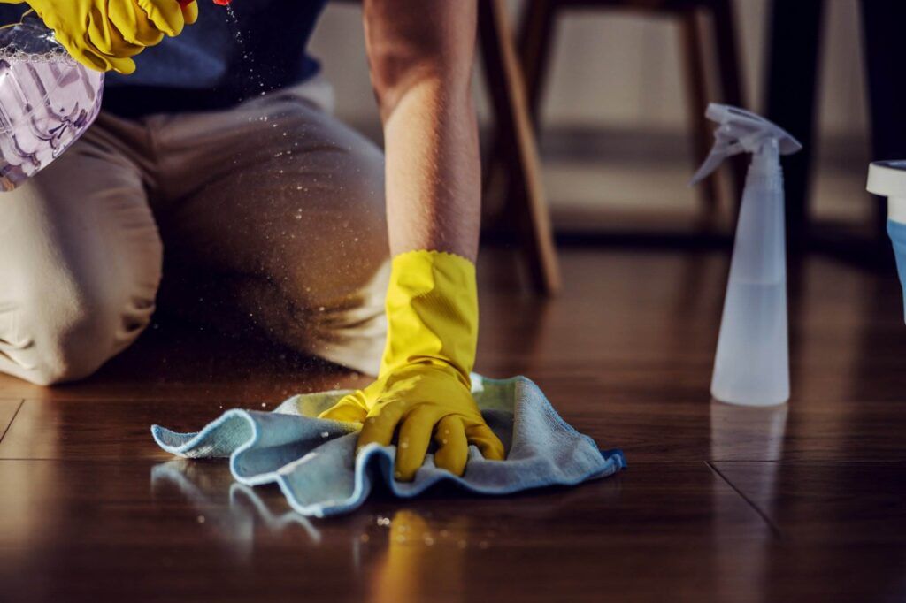 A person wearing yellow gloves diligently cleans a wooden floor with a cloth and spray bottle, enhancing the quality of life in their San Carlos home.