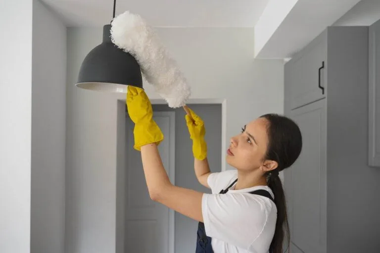 A person wearing yellow gloves and a white shirt cleans a black hanging light fixture in a San Carlos kitchen with a dusting tool, showcasing the meticulous attention to detail provided by Master Clean’s professional room cleaning services.