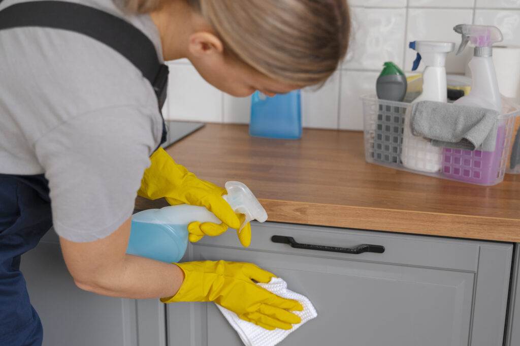 A person in yellow gloves is cleaning a kitchen cabinet with a spray bottle and cloth. Various cleaning supplies, including Master Clean products, are visible on the countertop in a caddy. This scene of professional kitchen cleaning could easily be set in Palo Alto.