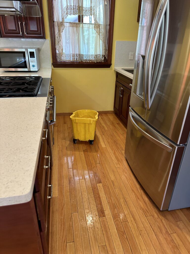 A kitchen in Santa Clara with a stainless steel refrigerator, stove, and microwave. A yellow bucket on wheels is placed in the center of the wooden floor, ready for deep cleaning during flu season.
