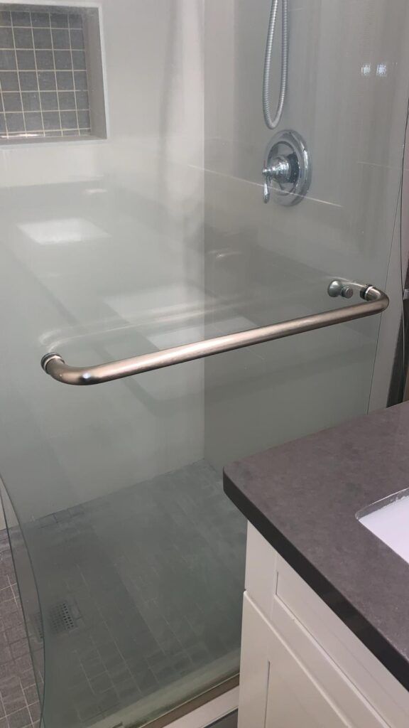 A glass shower enclosure with a stainless steel handle and a modern showerhead takes center stage, next to a bathroom counter and sink, ideal for thorough deep cleaning during flu season. 