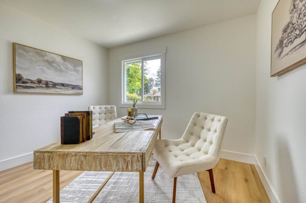 A minimalist home office with a wooden desk, two tufted chairs, books, and a gold desk lamp. The room has a window with a view of trees and two landscape paintings on the white walls.