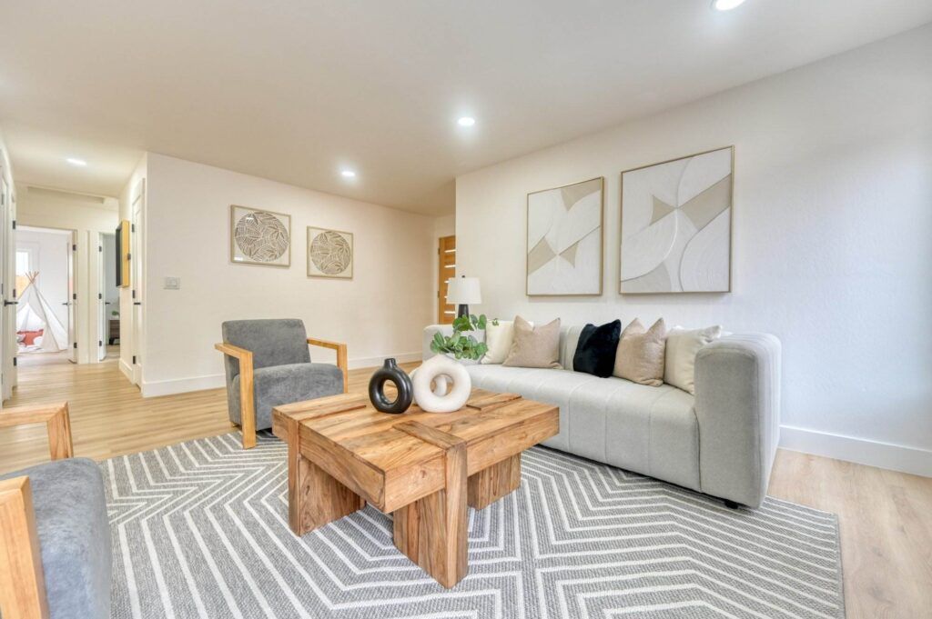 Modern living room with a grey sofa, wooden coffee table, two grey armchairs, gallery wall art, and a patterned rug. Room is bright with light wooden floors and recessed lighting.