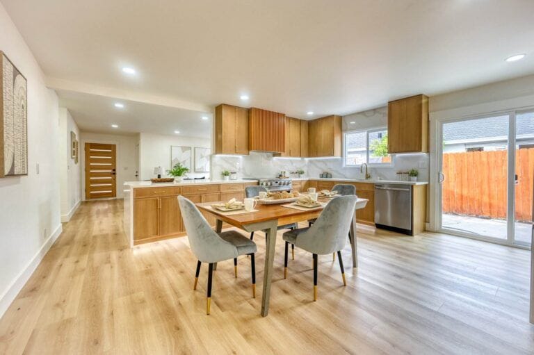 A modern kitchen and dining area featuring wooden cabinets, a dining table set for four, light wood flooring, and large sliding doors providing natural light.