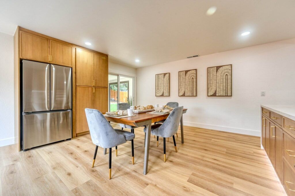 A modern dining area with a wooden table set with four gray upholstered chairs, a stainless steel refrigerator, light wooden cabinets, wall art, and large windows. The floor is light wood.