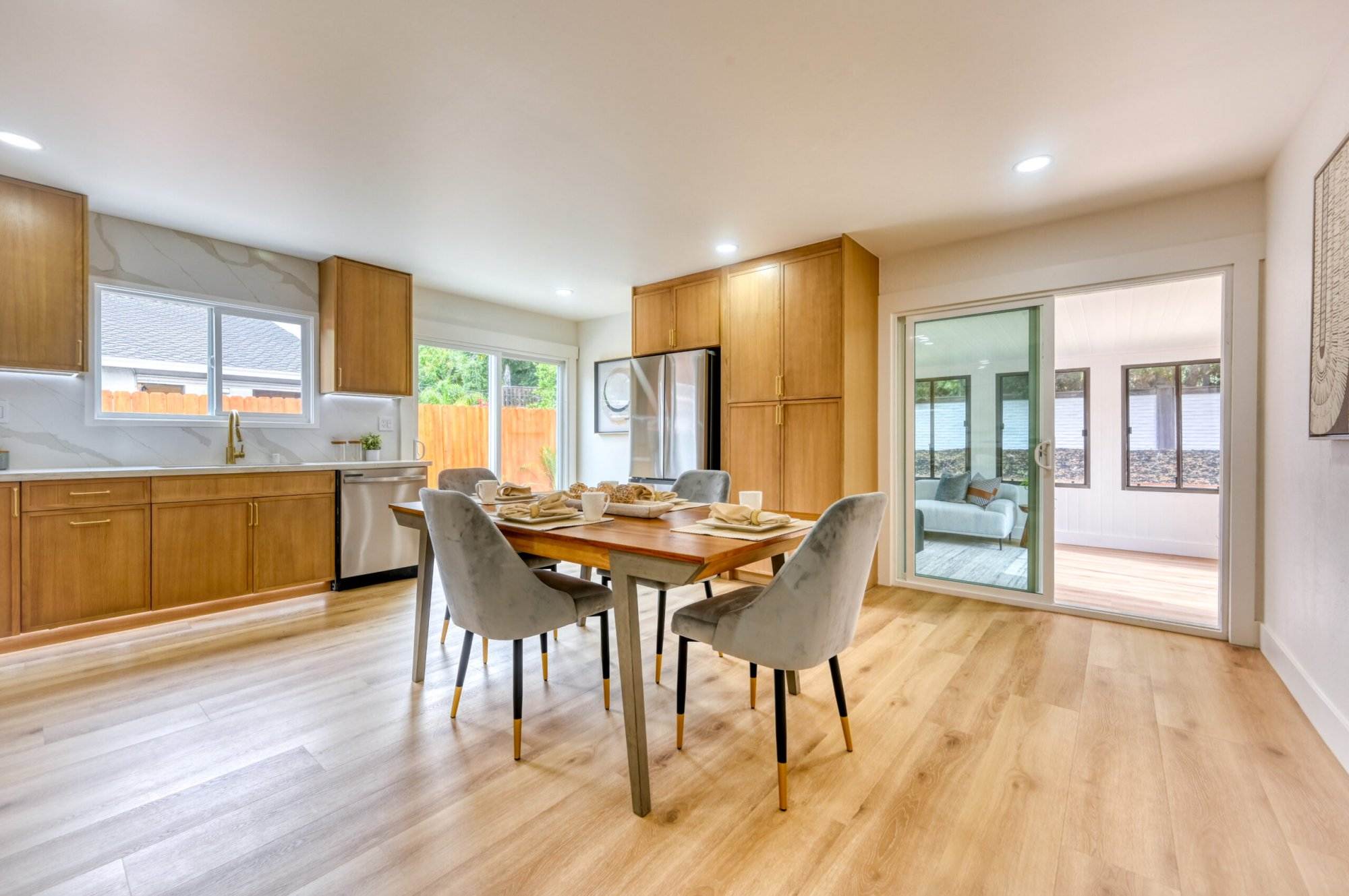 A bright, spacious kitchen and dining area featuring wooden cabinets, a wooden dining table, gray chairs, and sliding glass doors leading to an enclosed patio. Large windows allow natural light in.