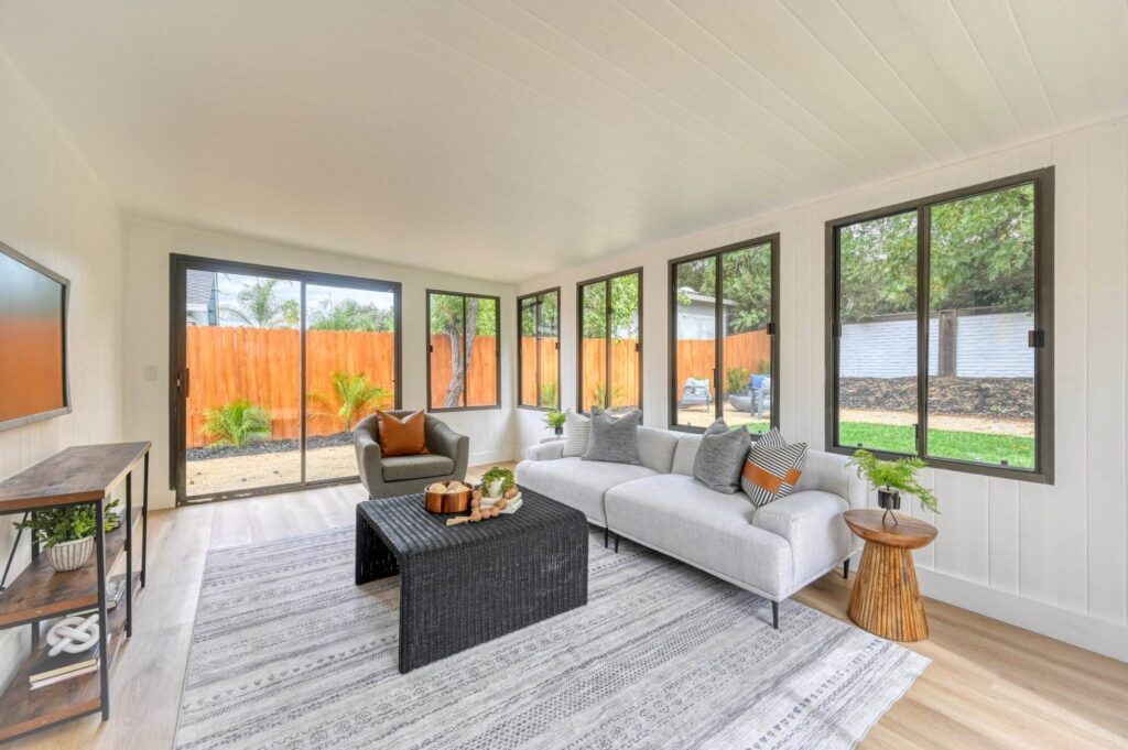 A bright sunroom with large windows, neutral walls and ceiling, gray and white furniture, a wicker coffee table, and a light gray area rug. A wooden side table and outdoor views are visible.