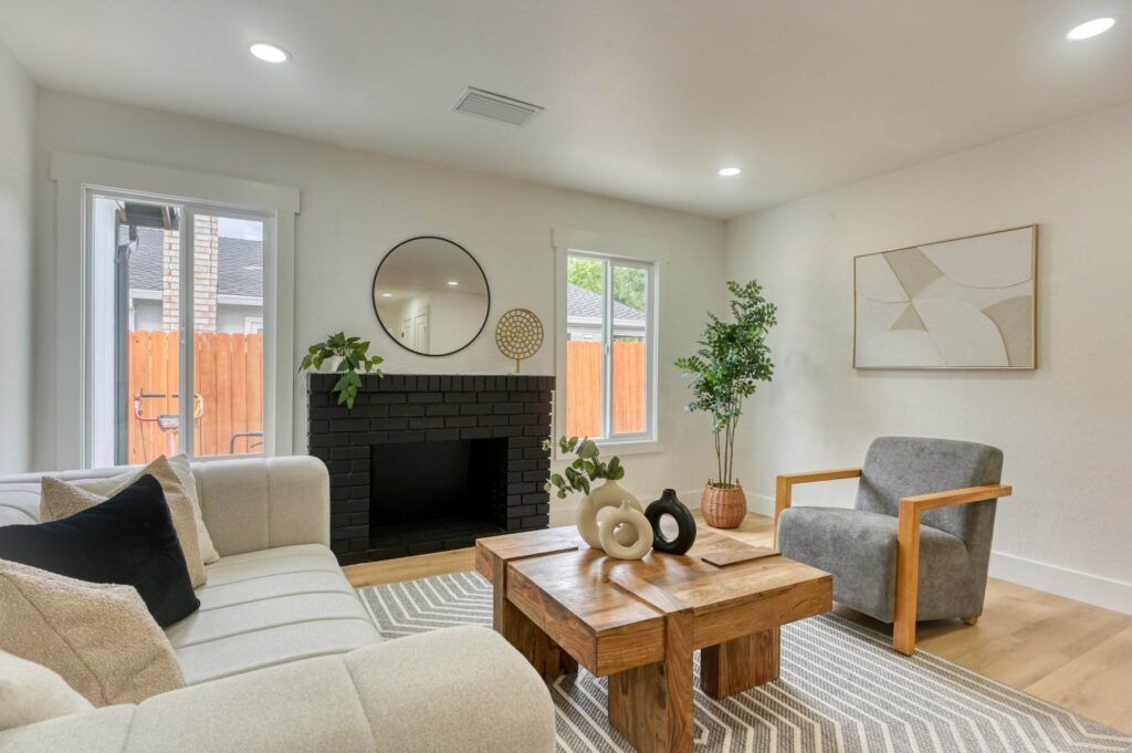 A modern living room with a beige couch, wooden coffee table, grey armchair, and black brick fireplace. The room features neutral decor, a round wall mirror, and abstract art on the wall.