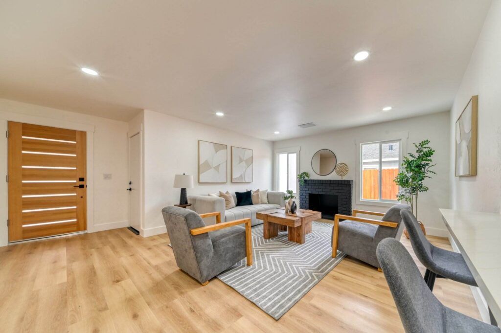 Modern living room with wooden flooring, gray cushioned chairs, a wooden coffee table, geometric decor on walls, and a small fireplace. Large windows bring in natural light.