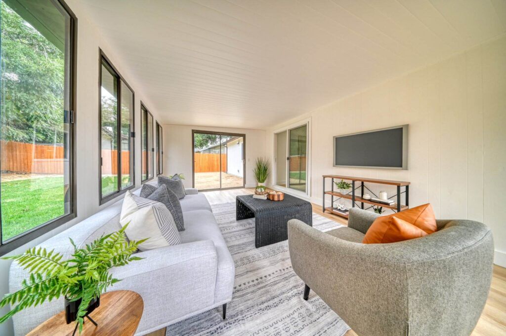 A sunroom with multiple windows, featuring a gray sofa, a gray armchair, a black coffee table, a TV on a stand, a small plant, and a view of the backyard.