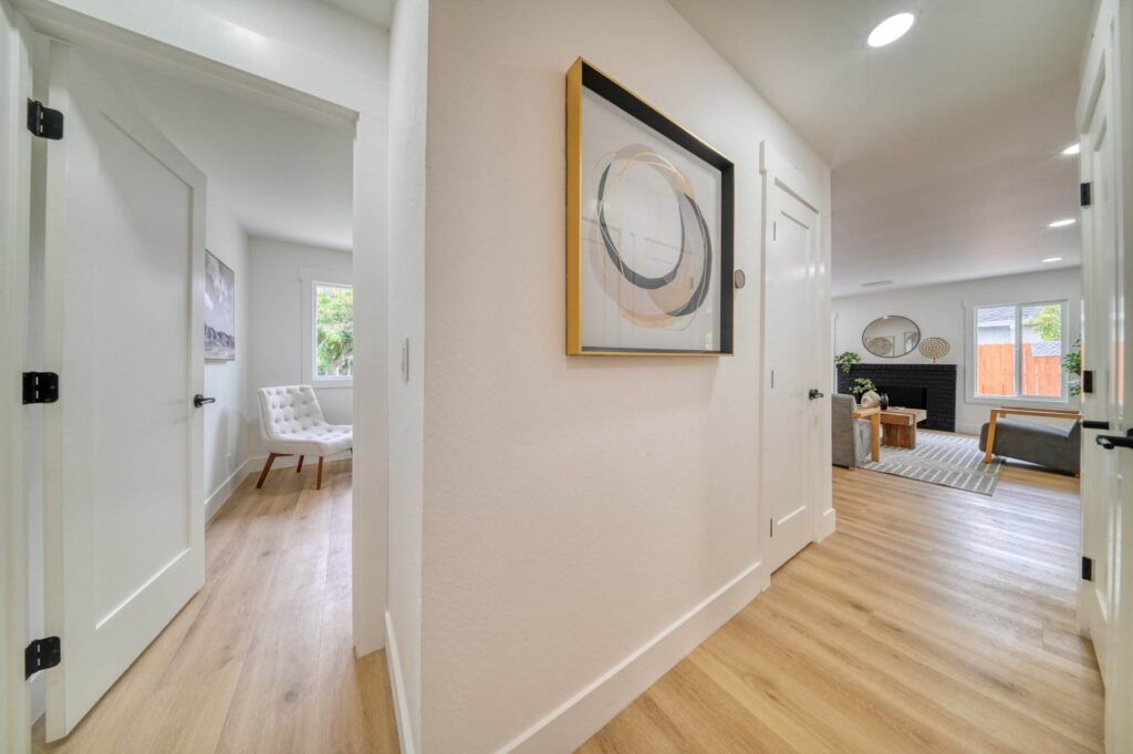 A hallway with light wood floors leads to multiple rooms. It features white walls, a piece of abstract wall art, and doors to a bedroom, a living area, and a possibly a bathroom.