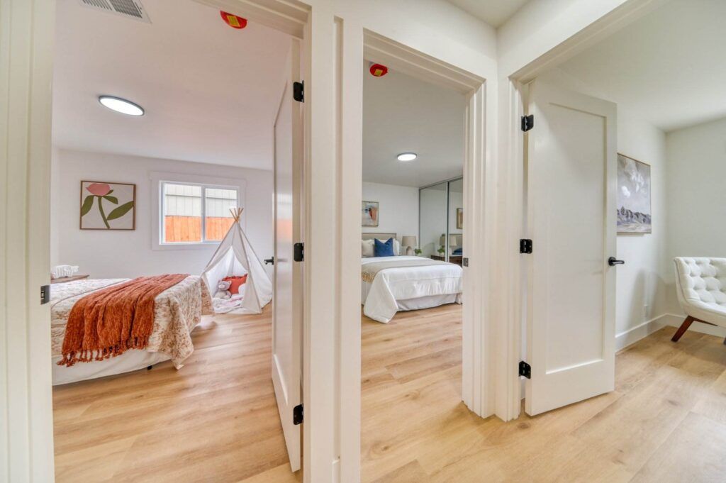 A hallway with two partially open doors revealing two bedrooms, each with double beds, light wood flooring, and white walls. The left room also has child-sized teepee.