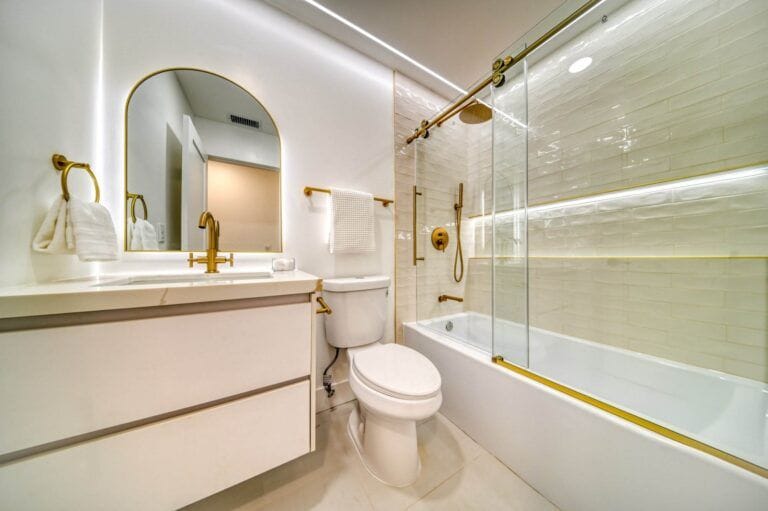 Modern bathroom features a vanity with mirror, a toilet, and a bathtub with glass doors. The fixtures are gold, complementing the white tiles and walls. Warm lighting illuminates the space.