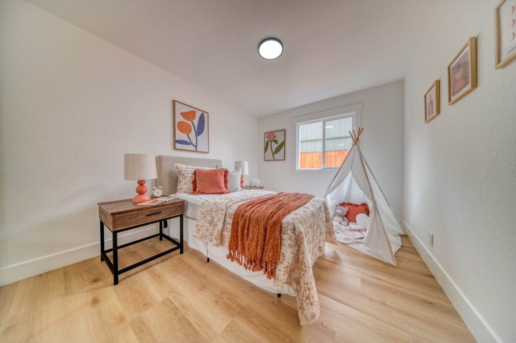 A cozy bedroom with a twin bed, orange blankets, a nightstand with a lamp, a small indoor tent, and framed wall art. The room has wooden flooring and a window letting in natural light.
