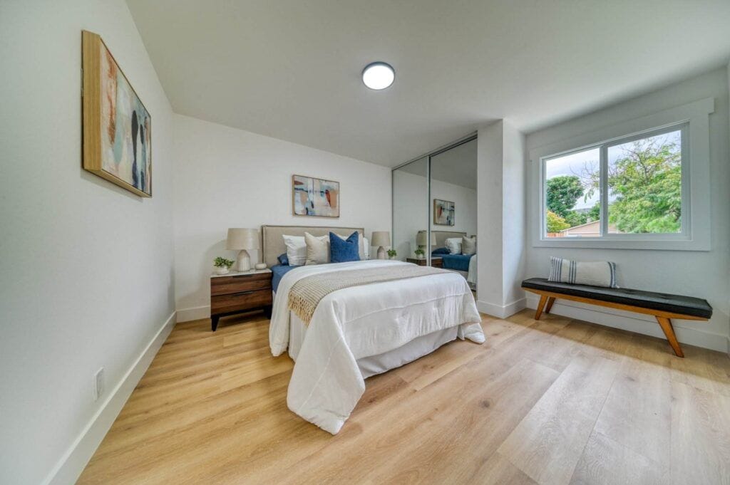 A modern bedroom with a bed, white bedding, two bedside tables with lamps, a bench by the window, and a mirrored closet. The room features wooden flooring and abstract wall art.