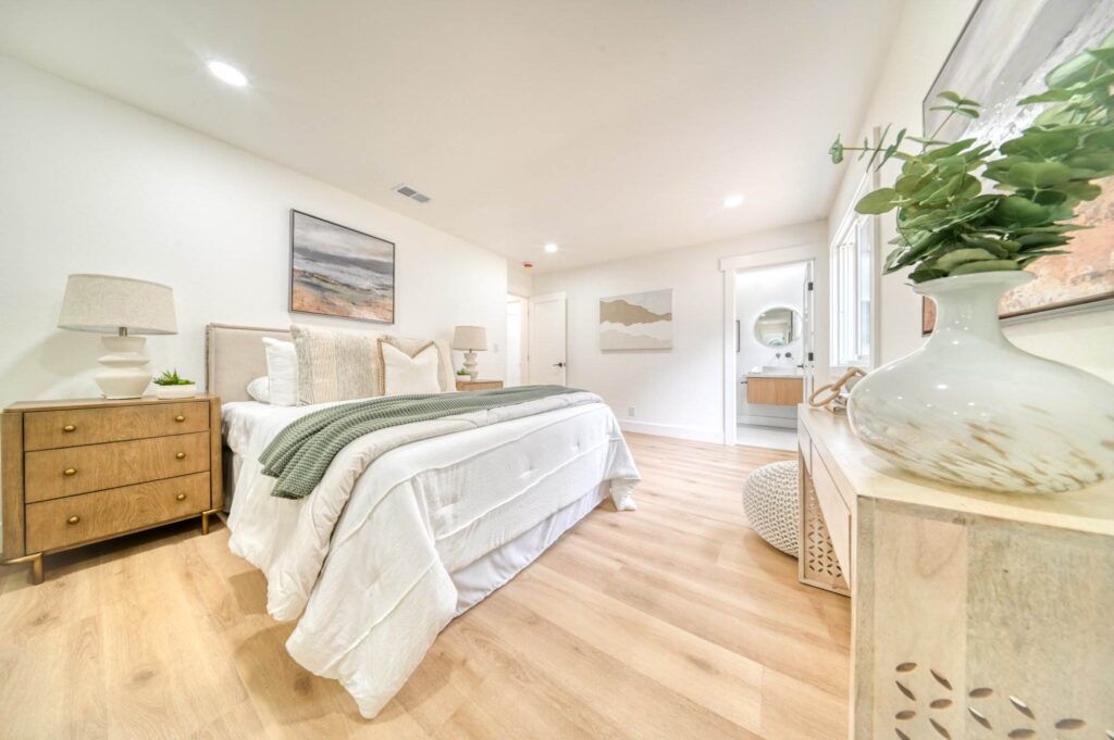 A modern bedroom with wooden flooring features a neatly made bed with white linens, green throw, and bedside tables with lamps. A large window and a dresser with a decorative vase are also visible.