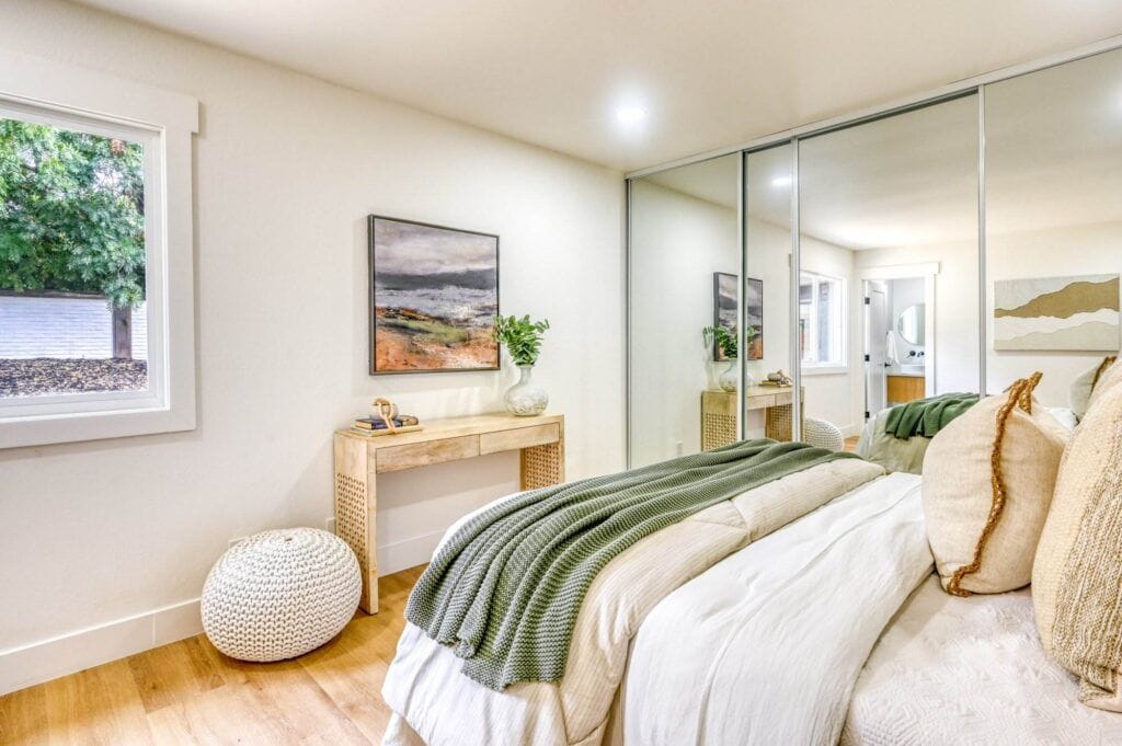 A cozy bedroom features a neatly made bed with green and beige accents, a light wood bedside table, a pouf, wall artwork, and a large mirrored closet. A window provides natural light.