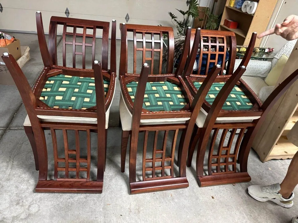 Four wooden chairs with green and beige woven seats are stacked upside down in a garage. A person's arm points at the chairs from the right side of the image.