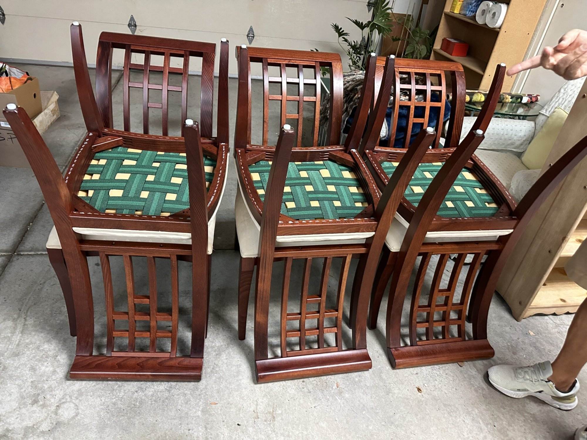 Four wooden chairs with green and beige woven seats are stacked upside down in a garage. A person's arm points at the chairs from the right side of the image.