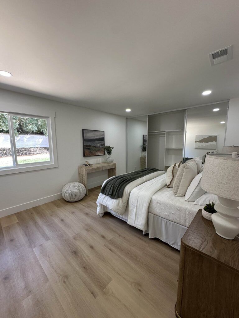 A modern bedroom with a double bed, wooden floor, minimalist decor, a window, a small table, and a mirrored sliding closet door. A lamp sits on a nightstand beside the bed.