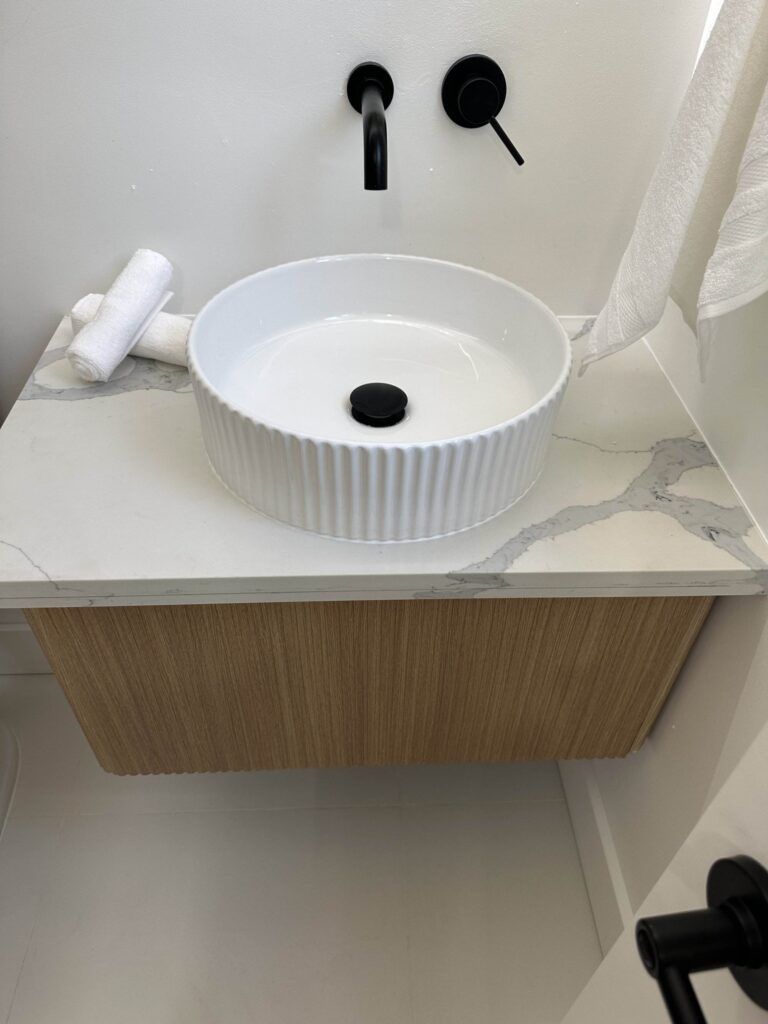 Modern bathroom sink with a ribbed white basin, black faucet, and black drain cover. Two rolled white towels are positioned on the left side of the countertop, which is mounted on a wooden cabinet.