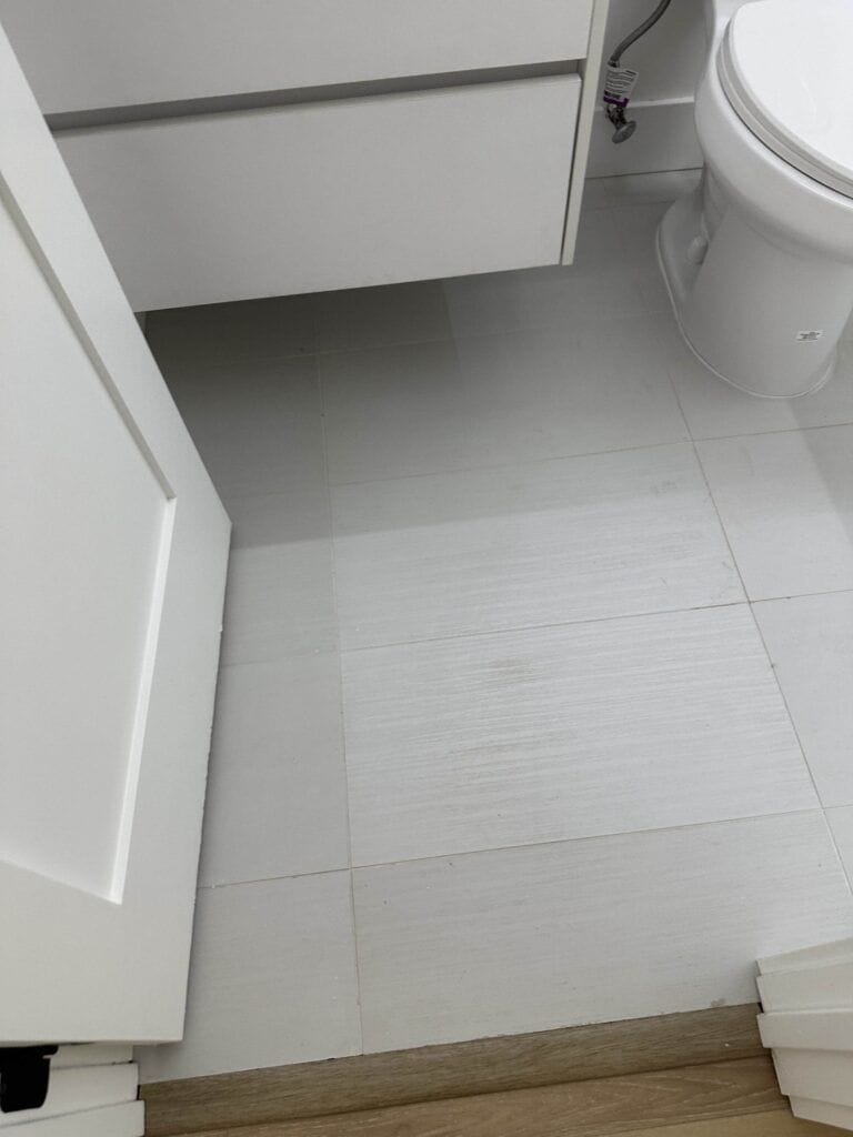 A bathroom with white floor tiles, a partial view of a closed cabinet, and a white toilet bowl on the right.
