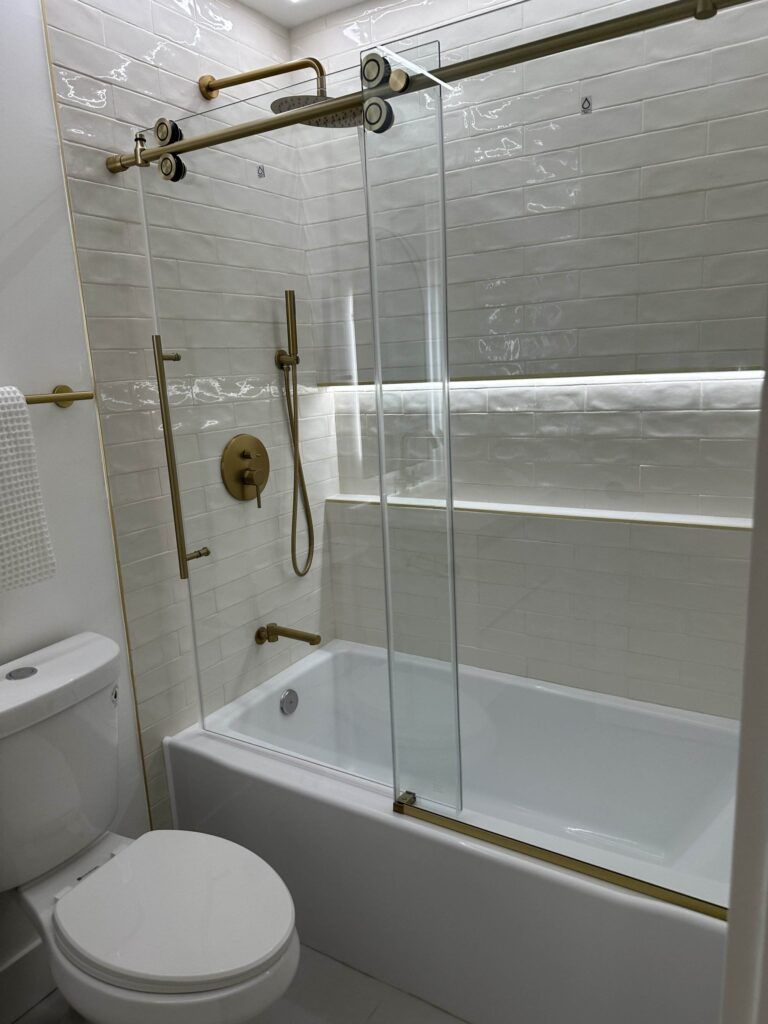 A modern bathroom featuring a white bathtub with a glass sliding door, gold fixtures, white tiled walls, and a towel on a rail next to a toilet.