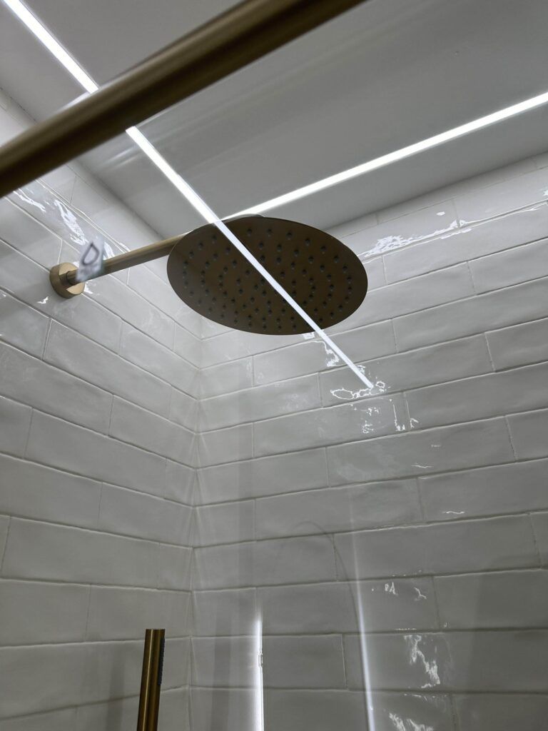 A modern shower with a round, wall-mounted rainfall showerhead against a white tiled wall, illuminated by overhead lighting.