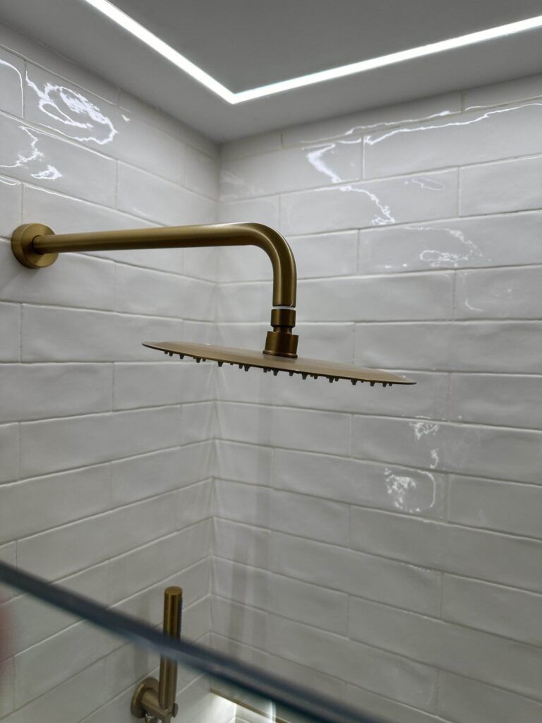 A square brass rainfall showerhead is mounted on a tiled wall in a shower with white subway tiles.