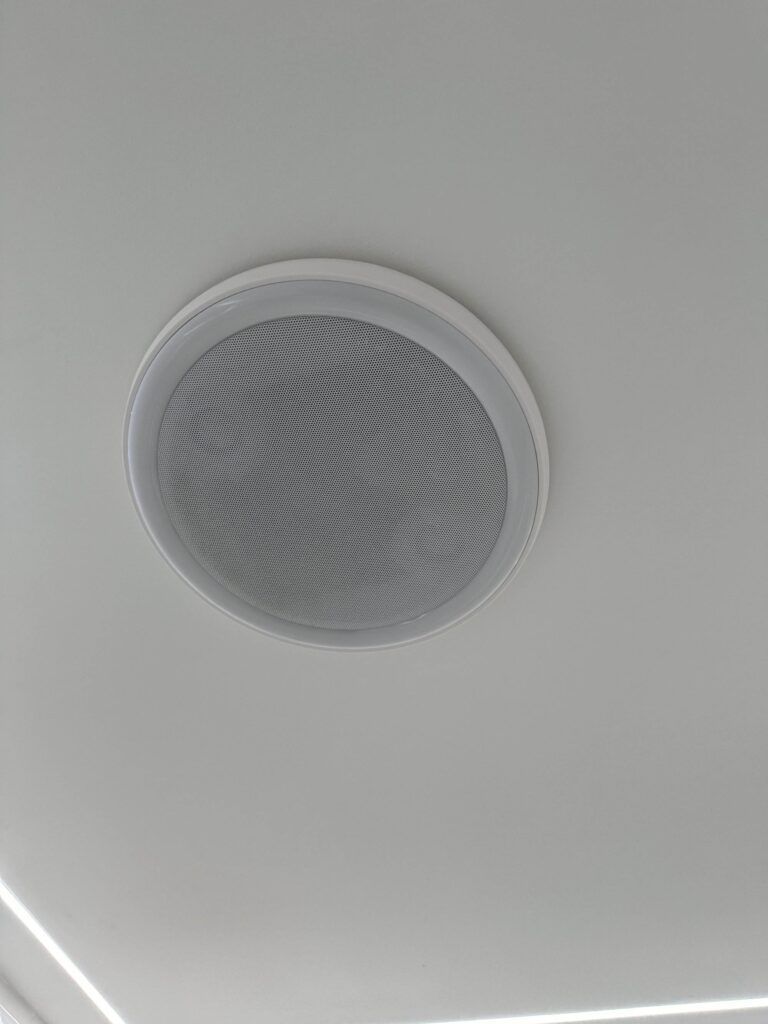 Ceiling-mounted round speaker with a white mesh cover in the center of a white ceiling.