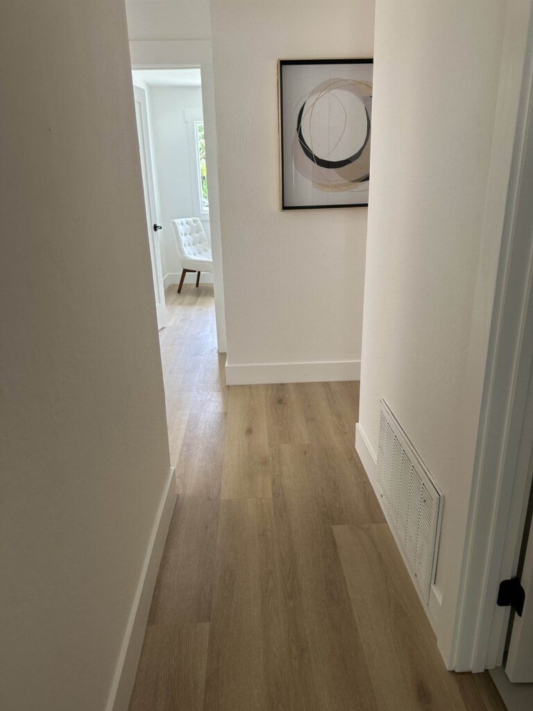 A well-lit hallway with wooden flooring leads to a room with a white chair near a window. A modern abstract painting hangs on the right wall.