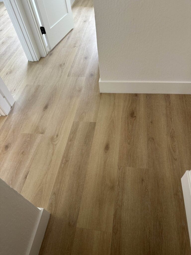 A hallway with light wood laminate flooring and white baseboards leads to an open door on the left, revealing more of the same flooring in the adjacent room.