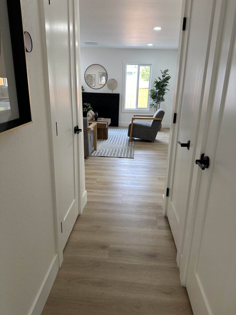 View of a modern living room with light wood flooring, seen from a hallway. The room has a gray sofa, a beige rug, a potted plant, a coffee table, and a wall-mounted mirror.