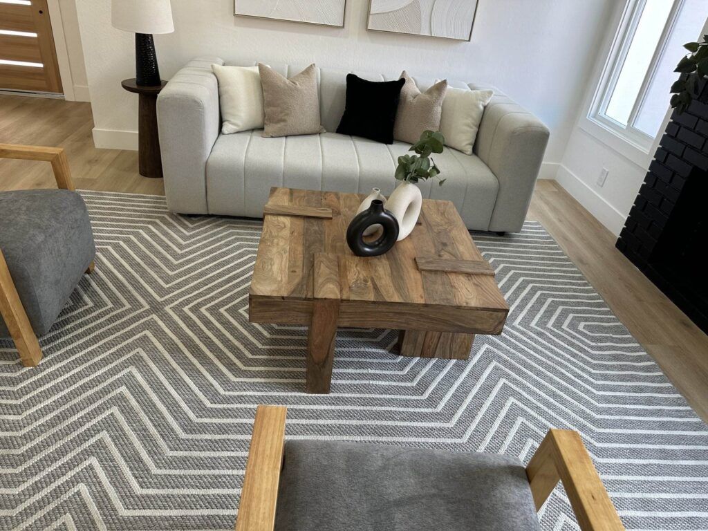 A modern living room features a gray couch with assorted pillows, a wooden coffee table, a chevron-patterned area rug, and two wooden-framed chairs. Decorative items sit on the table.