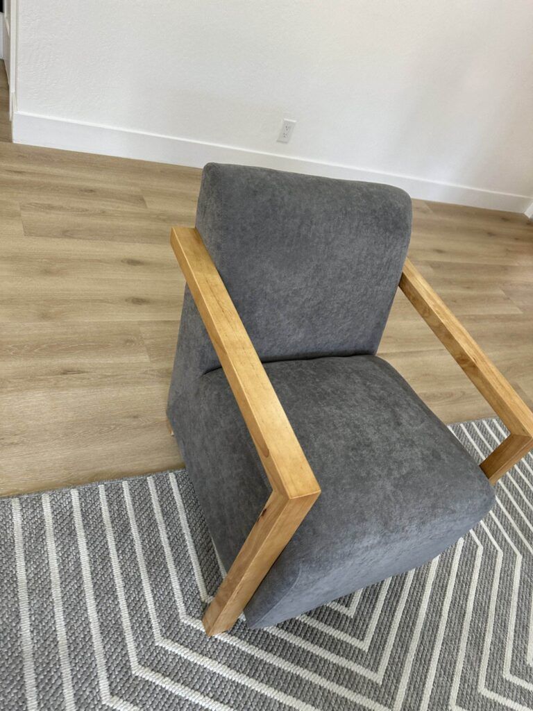 A gray cushioned chair with wooden armrests sits on a gray-striped rug over a wooden floor in a minimally furnished room.