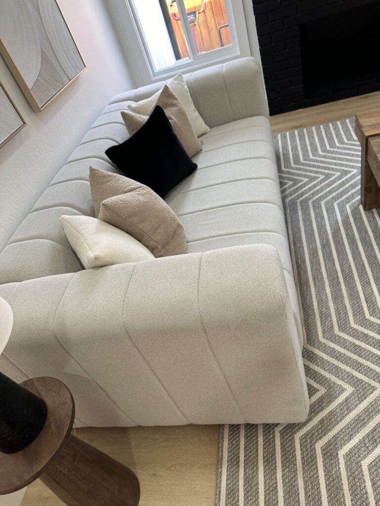 A light beige upholstered sofa with five pillows, two beige, one black, and two ivory, is placed near a black fireplace and a wooden coffee table on a gray and white patterned rug.