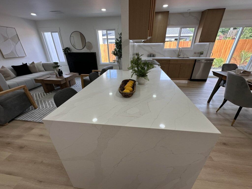 Modern open-concept kitchen and living space with a white marble island, wooden cabinets, light-colored floors, and minimalistic decor.