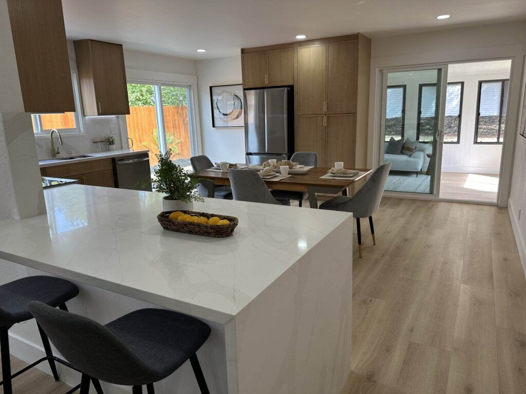 Modern kitchen and dining area with marble countertops, wooden cabinets, and a table set for six. Bar stools are positioned at the island, and large windows allow natural light to fill the space.