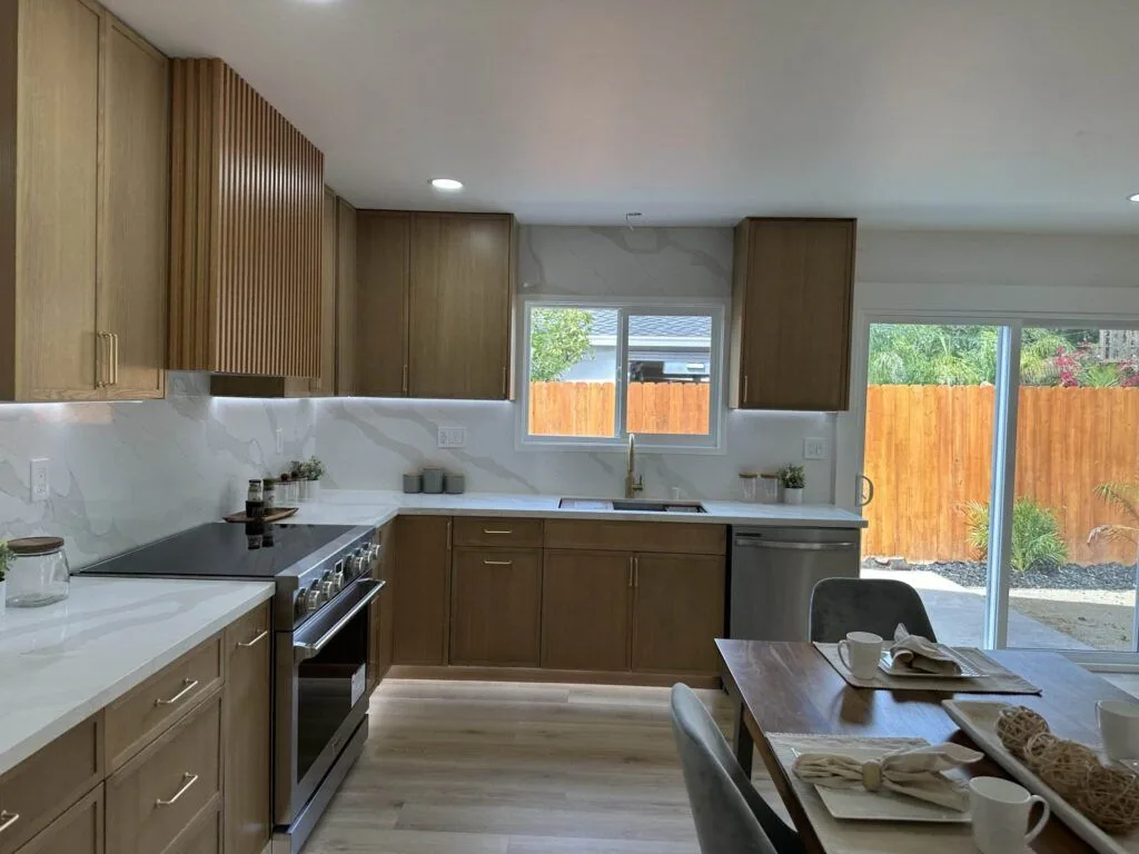 Modern kitchen with light wood cabinets, marble countertops and backsplash, stainless steel appliances, and a dining table. A sliding glass door opens to a backyard with a wooden fence.
