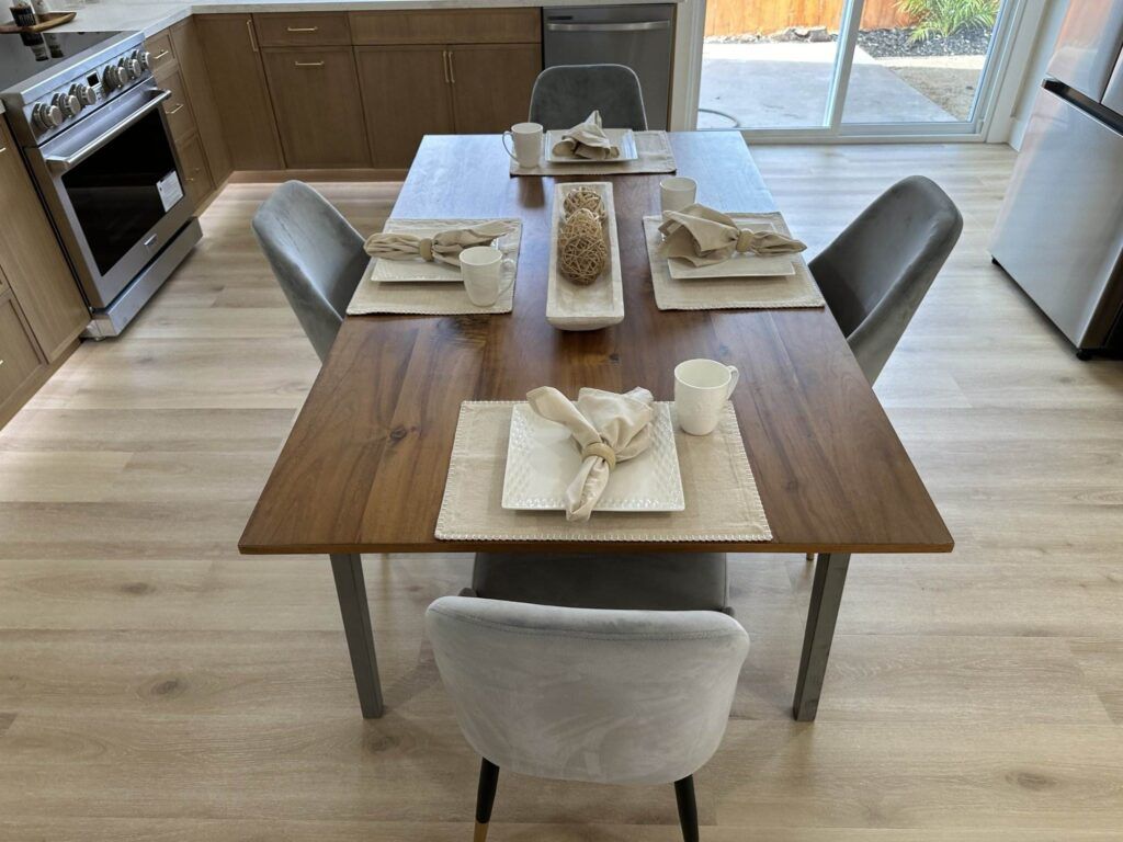 A dining table set for four with beige placemats, folded napkins, white cups, and a rectangular centerpiece in a modern kitchen with wooden flooring and stainless steel appliances.